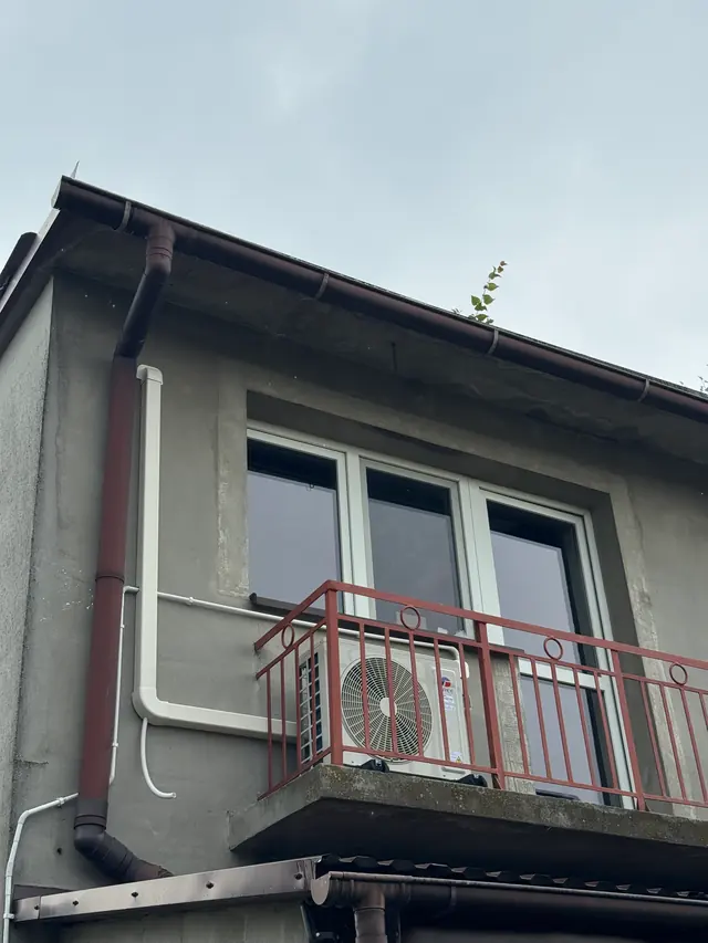Jednostka zewnętrzna na balkonie domu, przewody w białej listwie przy rynnie – Zadroże